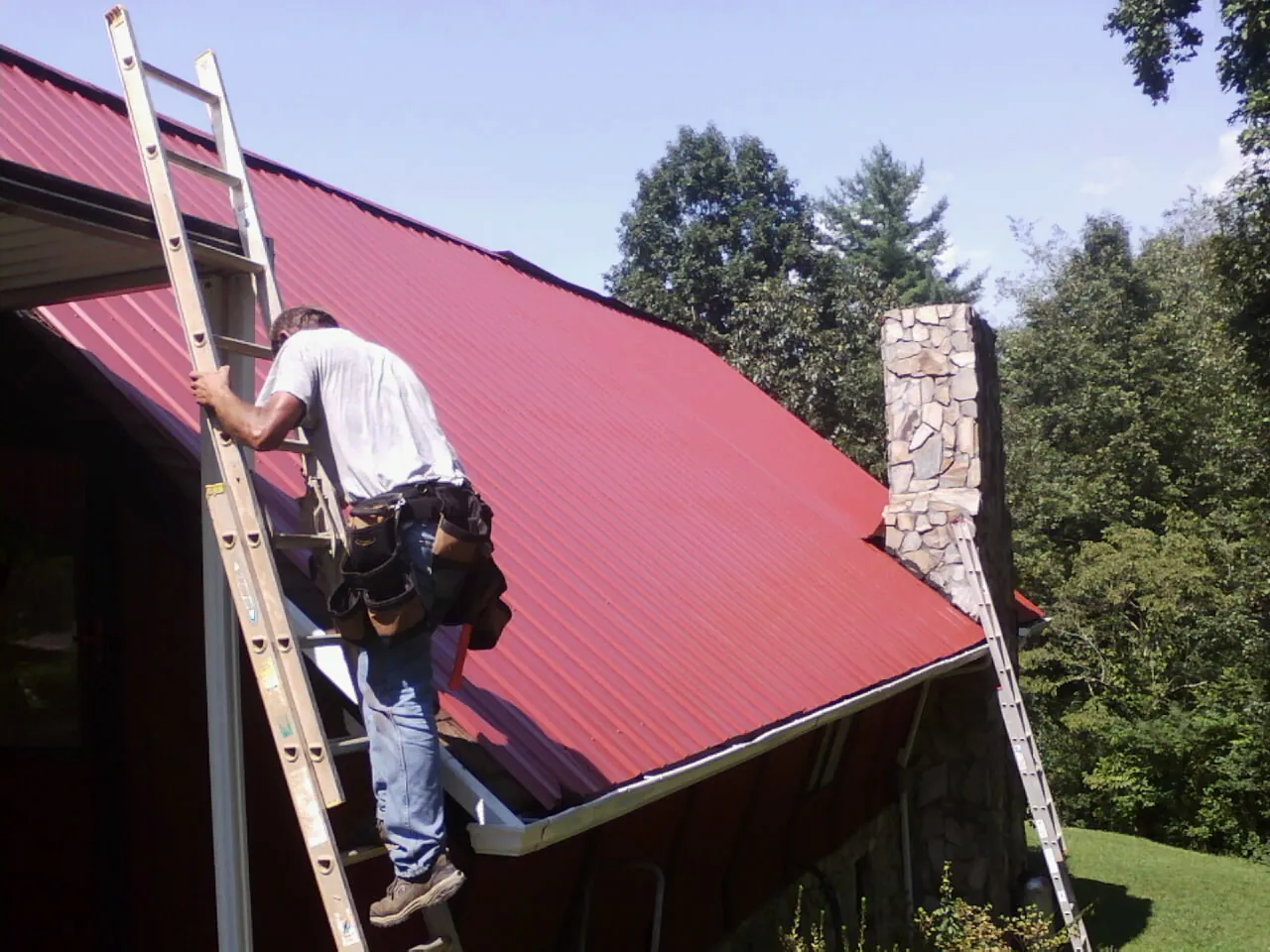 Roofer on a ladder beside a red metal roof and stone chimney — Soffit & Fascia Repair by Roofing Brothers in Byram